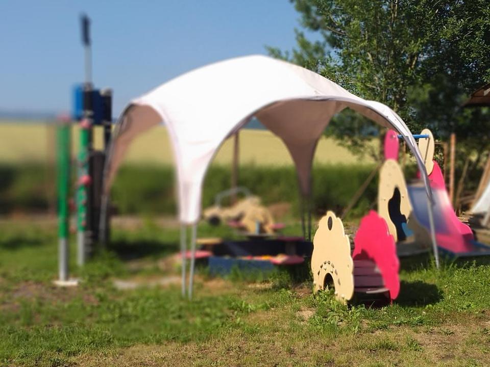Dětské hřiště v kempu — prolézačky, skluzavka a zastíněný altánek na trávě.